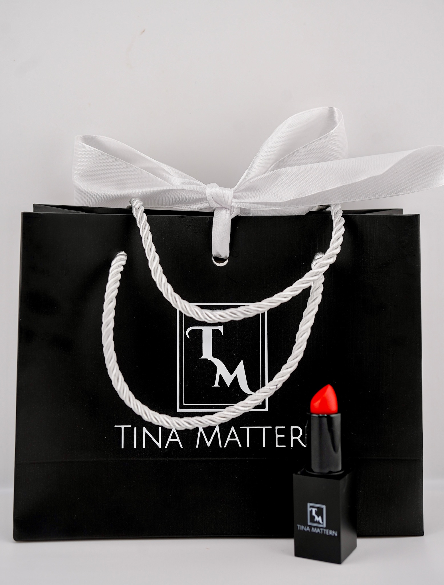 Black Tina Mattern gift bag with a white ribbon and a bold red lipstick in front.